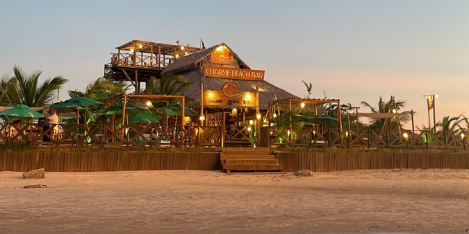 Fachada do Charme Beach Bar iluminada ao entardecer na praia em Atins, com estrutura de madeira, teto de palha e deck elevado.