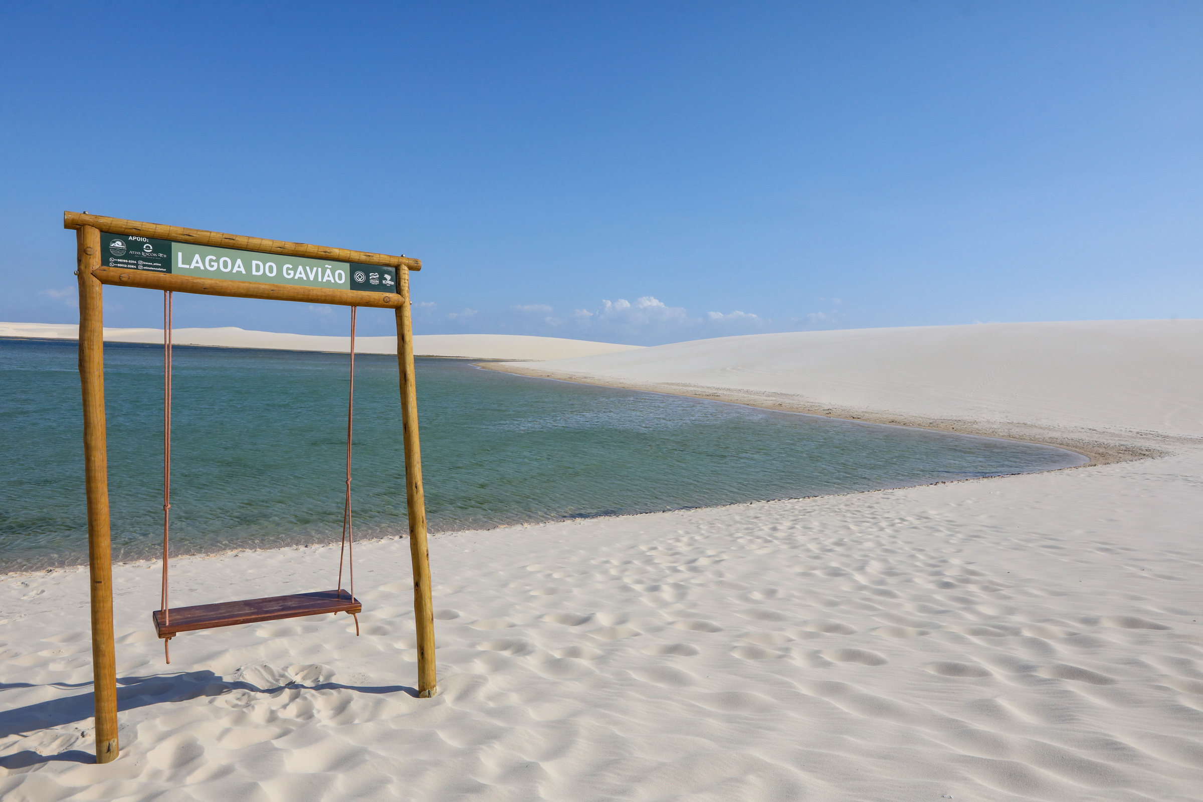 Wooden swing on the sand with the sign 'Lagoa do Gavião' facing the lagoon and dunes in Lençóis Maranhenses.