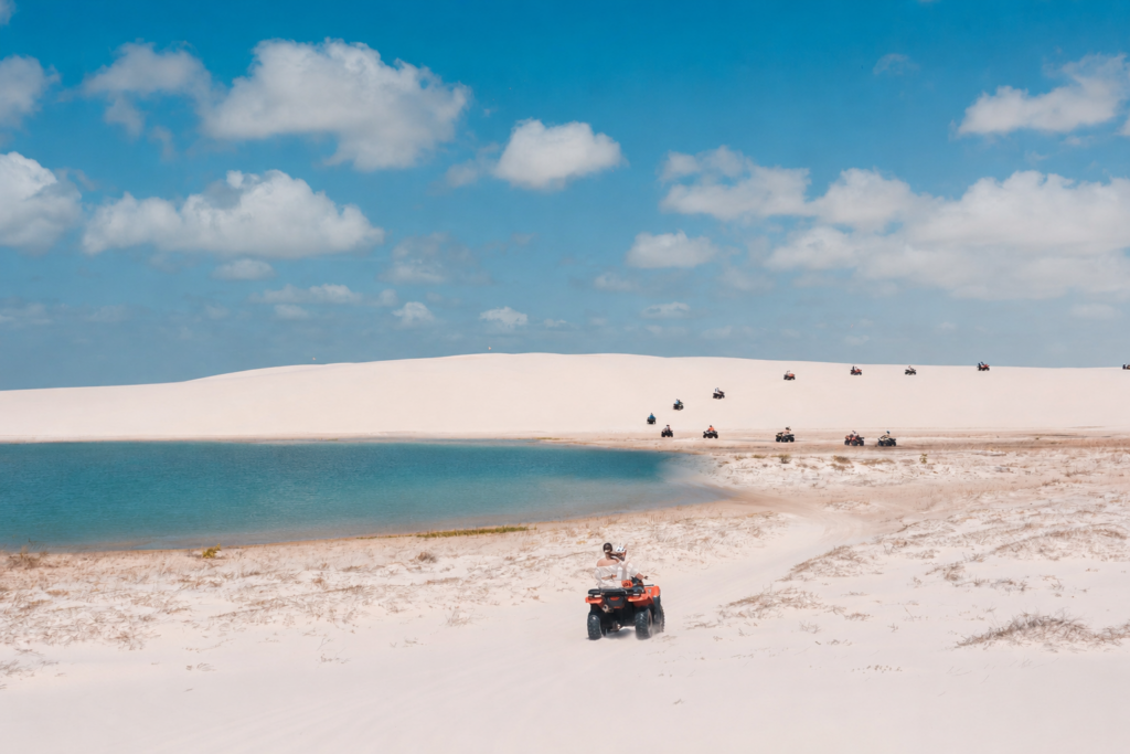 Pessoas pilotando quadriciclos nas dunas de areia branca ao lado de uma lagoa de águas azuis em Atins, Lençóis Maranhenses.