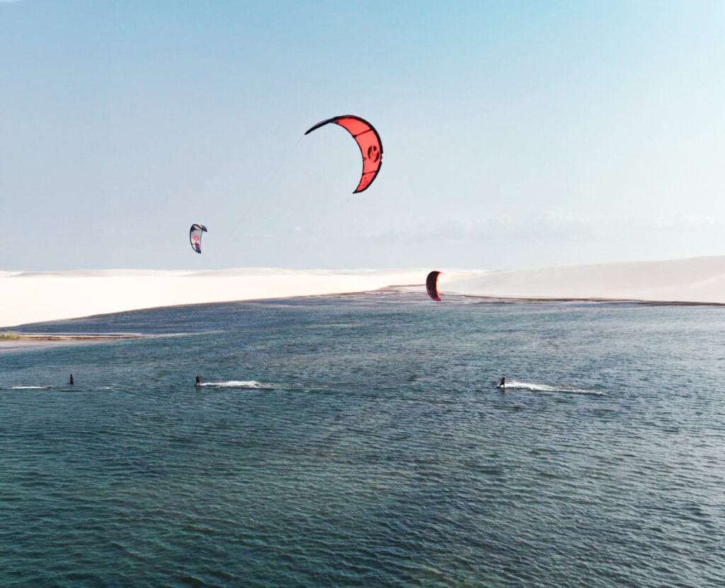 Vários kitesurfistas velejando em uma ampla lagoa nos Lençóis Maranhenses, com pipas coloridas no céu e dunas de areia branca ao fundo.