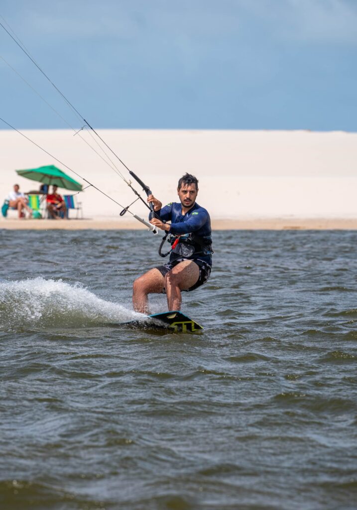Homem praticando kitesurf na água com dunas de areia branca ao fundo.