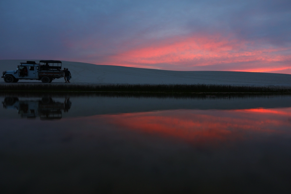 4x4 tour vehicle parked beside the dunes at dusk, with the reddish sunset sky reflected in the lagoon's water in Lençóis Maranhenses.