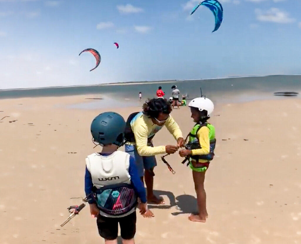 Instrutor de kitesurf ajustando o equipamento de duas crianças na praia, com pipas coloridas voando sobre a água ao fundo, em Atins.
