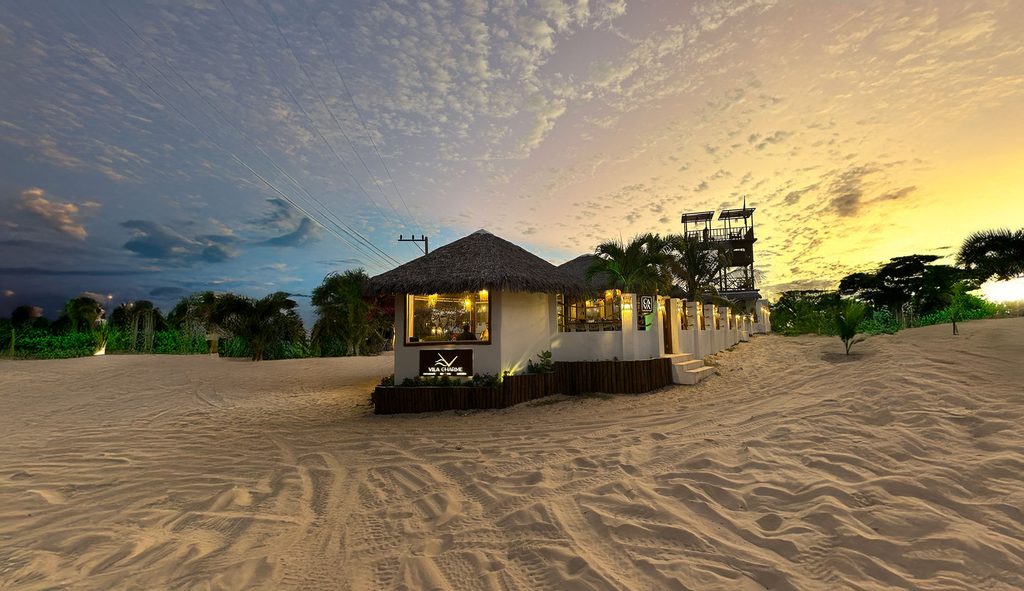 Vista exterior nocturna del restaurante Ça-Vá Charme Atins, una cabaña de madera y paja iluminada de forma cálida, situada sobre la arena de la playa de Atins con el cielo del atardecer al fondo.