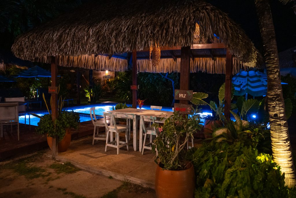 Área de comedor al aire libre del Mirante Charme Atins por la noche, con mesas y sillas de madera rústica bajo un techo de paja, iluminado con luces tenues y con plantas tropicales en primer plano y la piscina de fondo.