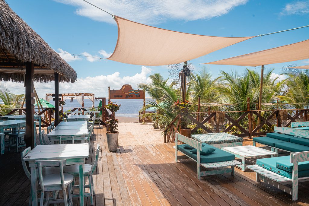 Terraza de madera del Atins Charme Beach Bar a la luz del día, con mesas y sillas de color blanco y azul claro, sombreada por lonas de tela y rodeada de vegetación tropical con vista al mar.